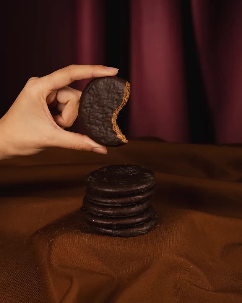 Una mano sostiene una galletita Malfatti bañada en chocolate negro, con un mordisco, sobre una pila de galletitas iguales.